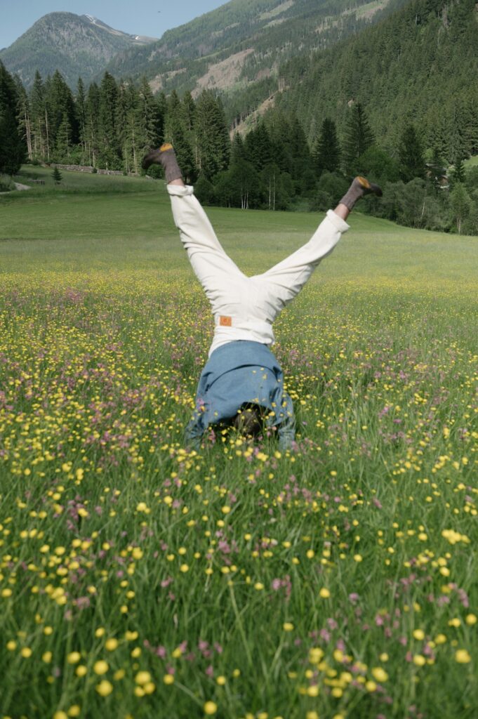 Person doing a headstand in a vibrant meadow filled with yellow and purple flowers, surrounded by lush green hills and forests in the background.