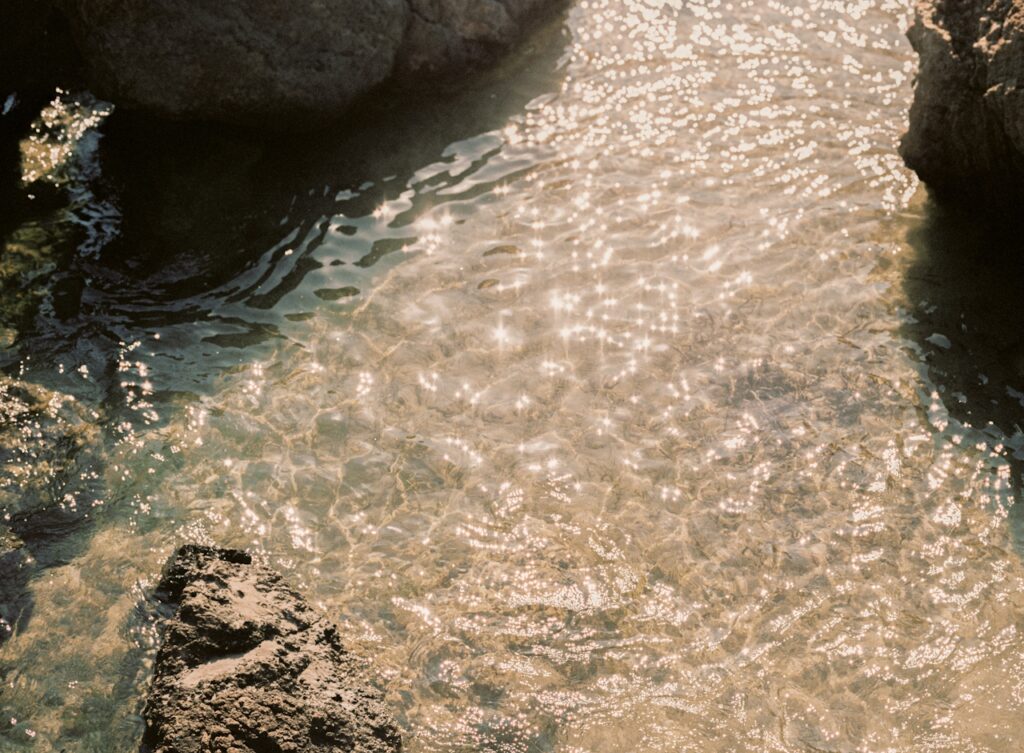 Close-up view of sunlit, clear water gently flowing over small pebbles, with sparkling light reflections and shadows cast by surrounding rocks.